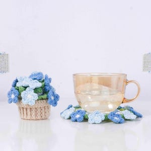 以下が含まれることがあります： 青と白の花で飾られたかぎ針編みのコースターの上に、取っ手付きの透明なガラスのティーカップが置かれています。左側には、青い花が入った小さなかぎ針編みの植木鉢があります。ポットはベージュです。