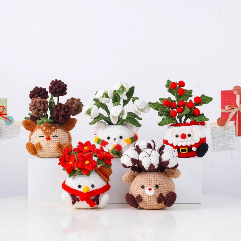 May include: A collection of five crocheted decorative planters, each shaped like a different animal or character. The planters are filled with various crocheted flowers and plants, including red berries and white blossoms. The characters include a reindeer, a snowman, and Santa Claus.