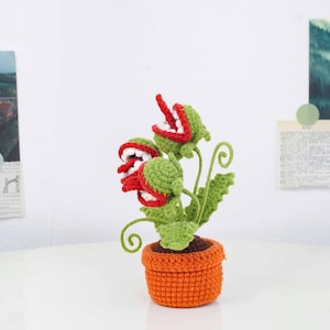 May include: A handmade crochet plant with three open-mouthed, red-lipped flowers with white teeth, set in a small orange pot. The green leaves and stems have a whimsical, cartoonish appearance, perfect for home decor.