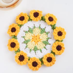 May include: A handmade crochet coaster featuring a sunflower design. The coaster has a white center with yellow, green, and brown accents. The outer edge is adorned with yellow sunflowers with brown centers and green leaves. The coaster is round and sits on a white surface.