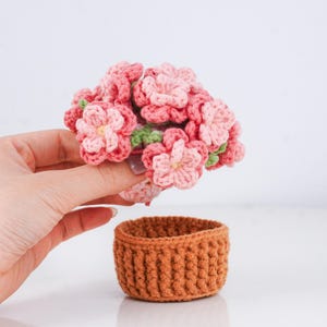 Puede incluir: Un conjunto de flores de ganchillo hechas a mano en tonos rosa y verde claro, sostenidas sobre una peque&ntilde;a cesta redonda de ganchillo marr&oacute;n. Las flores tienen una apariencia detallada y texturizada, y la cesta tiene un dise&ntilde;o acanalado.