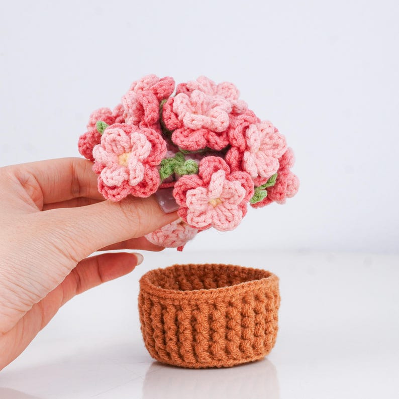 Puede incluir: Un conjunto de flores de ganchillo hechas a mano en tonos rosa y verde claro, sostenidas sobre una peque&ntilde;a cesta redonda de ganchillo marr&oacute;n. Las flores tienen una apariencia detallada y texturizada, y la cesta tiene un dise&ntilde;o acanalado.