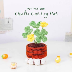 May include: A crocheted Oxalis Cat Leg Pot with green leaves and yellow flowers. The pot is brown and orange with white cat-paw feet. The text "PDF PATTERN Oxalis Cat Leg Pot" is at the top. Decorative items are scattered around the pot.