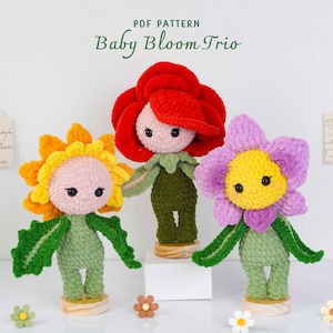 Könnte beinhalten: Drei handgefertigte gehäkelte Blumendolls. Die Puppen haben blütenförmige Köpfe in Gelb, Rot und Lila, mit grünen Körpern und Armen. Der Text "Baby Bloom Trio" befindet sich oben im Bild. Die Puppen stehen auf Holzsockeln.
