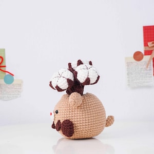 May include: A crocheted cotton plant in a tan pot with a brown stem and white cotton blooms. The pot is shaped like a small animal with brown accents and a red nose. The handmade item is on a white surface.