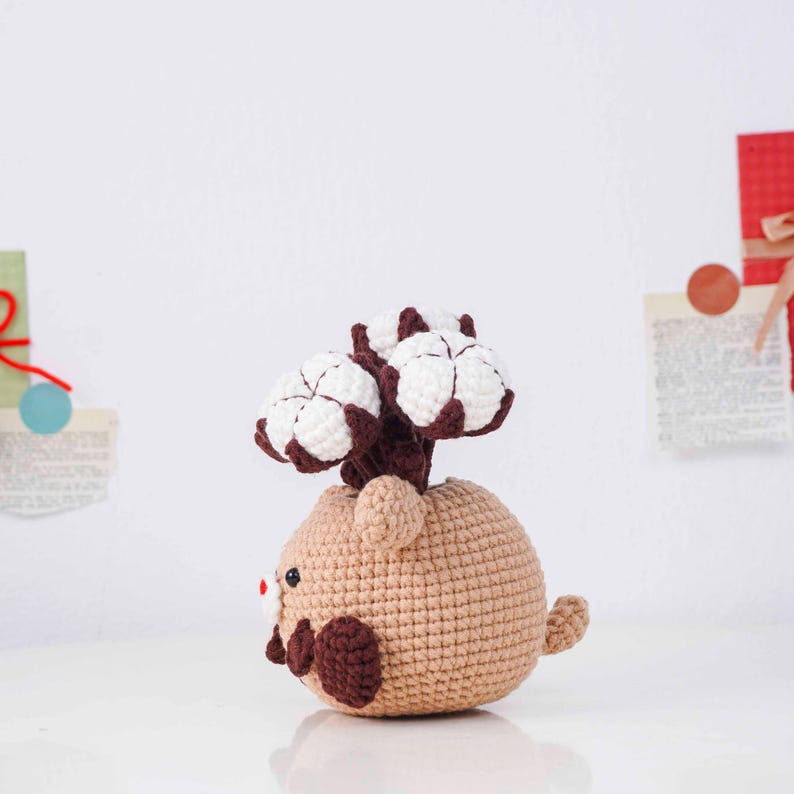 May include: A crocheted cotton plant in a tan pot with a brown stem and white cotton blooms. The pot is shaped like a small animal with brown accents and a red nose. The handmade item is on a white surface.