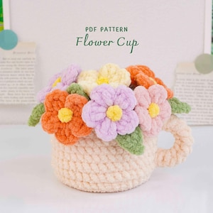 May include: A crochet cup in a light beige yarn filled with a bouquet of colorful yarn flowers in shades of orange, yellow, lavender, and pink, accented with green leaves. The text above the cup reads PDF PATTERN Flower Cup.