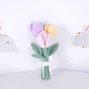 May include: A handcrafted crochet bouquet of tulips in pink, purple, and yellow. The flowers have green stems and leaves, tied with a white bow. The bouquet is set against a white backdrop.