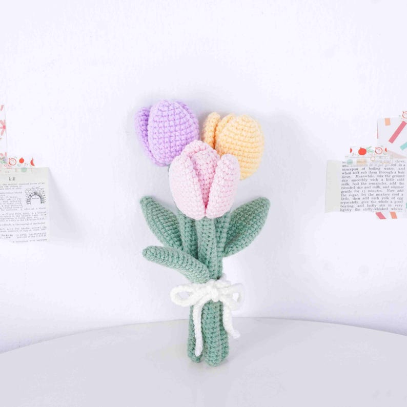 May include: A handcrafted crochet bouquet of tulips in pink, purple, and yellow. The flowers have green stems and leaves, tied with a white bow. The bouquet is set against a white backdrop.