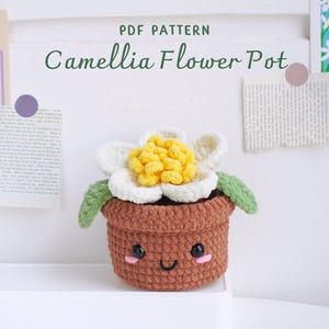 May include: A crocheted camellia flower pot with a smiling face. The pot is brown and the flower is white with a yellow center. The text "PDF PATTERN Camellia Flower Pot" is above the pot.