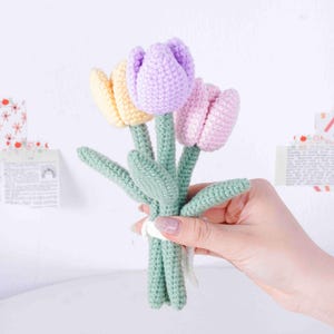 May include: A bunch of three crocheted tulips in pastel shades: yellow, lilac, and pink. The stems and leaves are green, and the flowers are tied with a white ribbon. The handmade flowers are held in a person's hand.