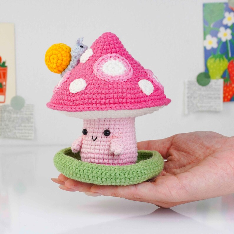 May include: A handmade crochet mushroom figure with a pink cap and white spots. The mushroom has a smiling face and arms, sitting in a green base. A yellow snail with a gray shell is on the cap. A whimsical, decorative item.