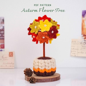 Puede incluir: Un árbol de flores de ganchillo con flores rojas, amarillas, naranjas y verdes sobre un tallo marrón. La base es una maceta cubierta con una funda de ganchillo texturizada en crema, naranja y marrón oscuro. El texto "PDF PATTERN Auturm Flower Tree" es visible.