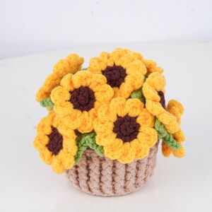 May include: A crocheted bouquet of sunflowers in a small, brown basket. The sunflowers have bright yellow petals and dark brown centers. Green leaves are visible between the flowers. The basket is a light brown color.
