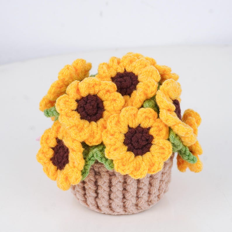 May include: A crocheted bouquet of sunflowers in a small, brown basket. The sunflowers have bright yellow petals and dark brown centers. Green leaves are visible between the flowers. The basket is a light brown color.