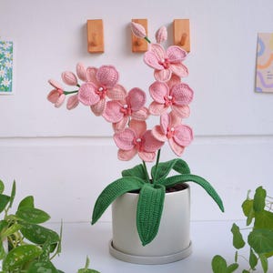 May include: A pink crocheted orchid plant in a white pot with green crocheted leaves. The plant is sitting on a white surface.