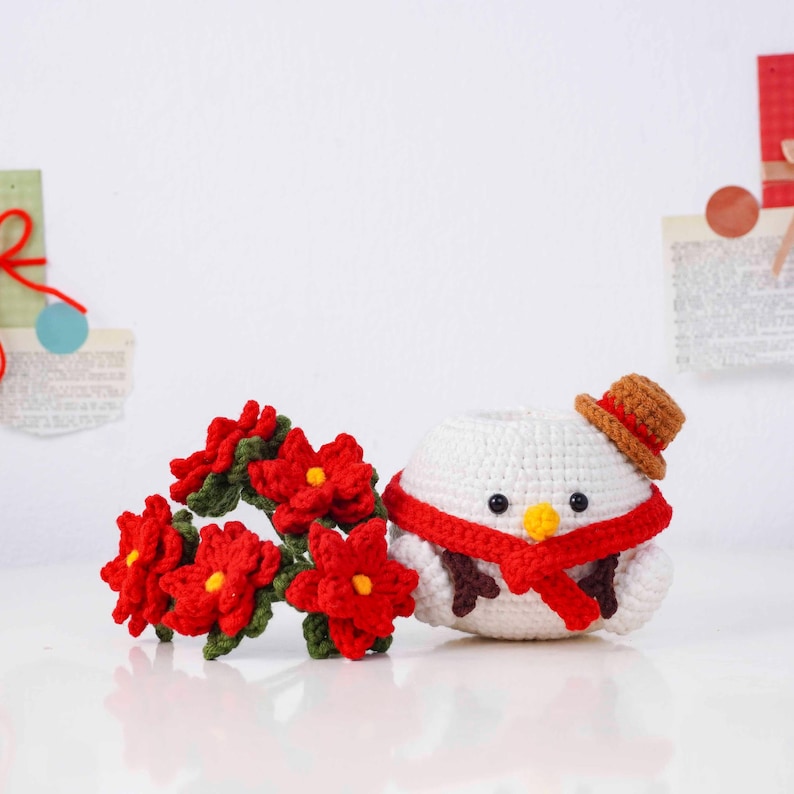 May include: A crocheted snowman with a brown hat and red scarf, next to a garland of red poinsettia flowers. The snowman has a yellow beak and brown twig arms. The items are handmade and festive.