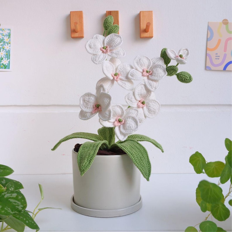 May include: A white crocheted orchid plant with pink centers in a gray ceramic pot. The plant has green crocheted leaves.