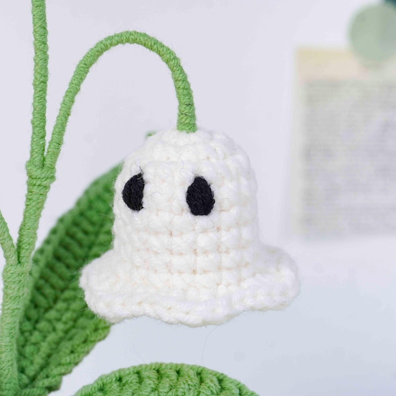 May include: A handmade crochet flower with a white bell-shaped bloom and two black eye-like details. The flower has a green stem and leaf, suggesting a craft project or decorative item.