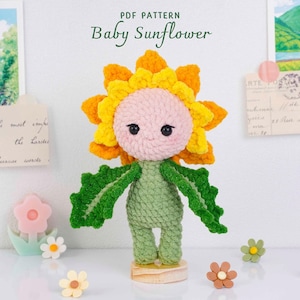 Könnte beinhalten: Gehäkelte Sonnenblumenpuppe mit gelb-orangem Blütenkopf, rosa Gesicht und grünem Stiel und Blättern. Die Puppe steht auf einem Holzsockel. Der Text "PDF PATTERN Baby Sunflower" steht oben.