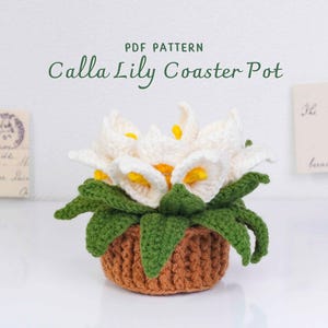 May include: A crocheted Calla Lily Coaster Pot. The pot is brown, with green leaves and white and yellow flowers. The text "PDF PATTERN Calla Lily Coaster Pot" is above the pot. There are vintage postcards in the background.