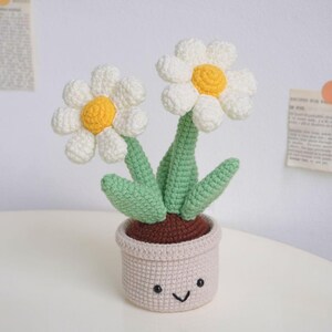 Puede incluir: Una planta de margarita de crochet con dos flores blancas y hojas verdes en una maceta beige con una base marr&oacute;n y una cara sonriente.