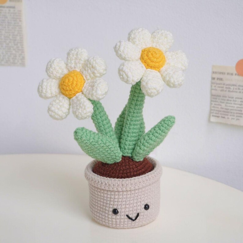 Puede incluir: Una planta de margarita de crochet con dos flores blancas y hojas verdes en una maceta beige con una base marr&oacute;n y una cara sonriente.