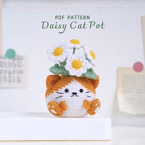 May include: Crochet pattern for a daisy cat pot. The pot is a white cat with orange ears and paws. The cat is wearing a pot with a bouquet of white daisies.