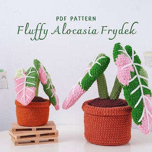 Puede incluir: Dos plantas de Alocasia Frydek tejidas a ganchillo en macetas naranjas. Las hojas son rosas, verdes y blancas. Las macetas están sobre un palé de madera. El texto "PDF PATTERN" y "Fluffy Alocasia Frydek" se muestran encima de las plantas.