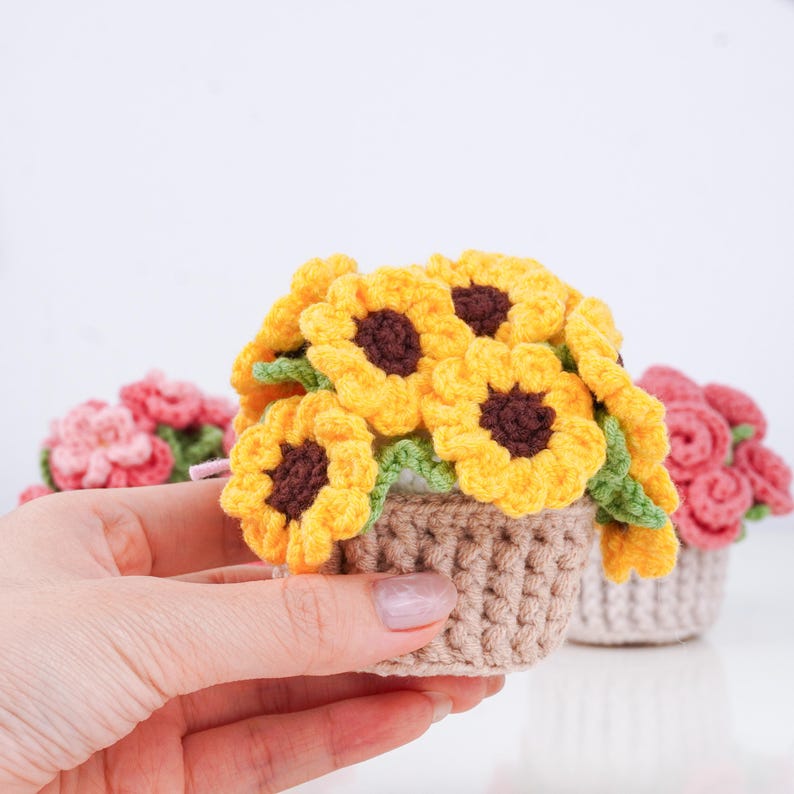 May include: A crocheted miniature sunflower arrangement in a small brown pot. The sunflowers are yellow with brown centers and green leaves. Other crocheted flower arrangements are in the background.