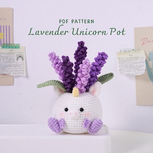 Op de afbeelding: Een gehaakte witte eenhoornpot met paarse lavendelbloemen. De eenhoorn heeft een gele hoorn, groene bladeren en paarse voetjes. De tekst "PDF PATTERN Lavender Unicorn Pot" staat boven de pot.