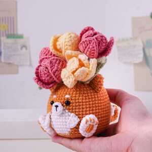 May include: A crocheted orange Shiba Inu dog with white accents, holding a bouquet of pink and yellow flowers. The handmade amigurumi features a cute design with a small tail and paw details. A unique, handcrafted gift.