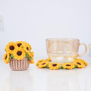 May include: A clear glass teacup with a handle sits on a crocheted sunflower coaster. To the left, a small crocheted basket overflows with yellow and brown crocheted sunflowers. The items are on a white surface.