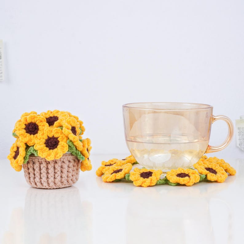 May include: A clear glass teacup with a handle sits on a crocheted sunflower coaster. To the left, a small crocheted basket overflows with yellow and brown crocheted sunflowers. The items are on a white surface.