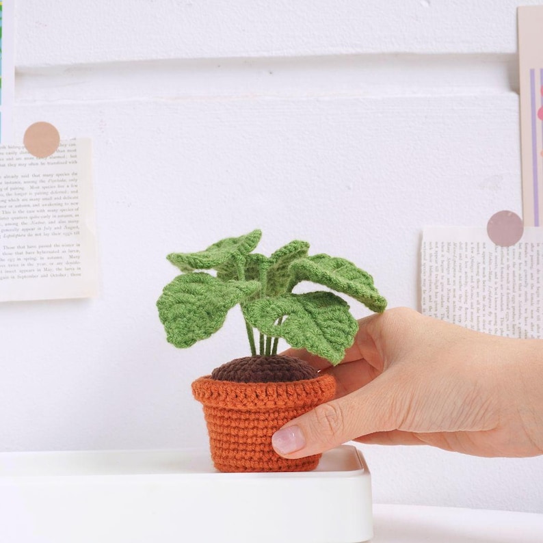 May include: A crocheted plant in a brown pot. The plant has green leaves and is sitting on a white surface.