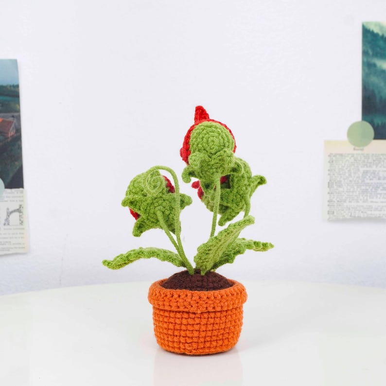 May include: A handmade crochet plant with red flowers and green leaves in an orange pot. The plant has a brown base and is set on a white surface. The crochet work is detailed and the colors are vibrant.