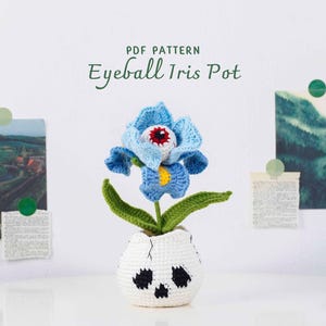 Op de afbeelding: Een gehaakte Eyeball Iris Pot, met een blauwe bloem met een rood en wit oogbalcentrum, groene bladeren en een witte schedelvormige pot met zwarte accenten. De tekst "PDF PATTERN Eyeball Iris Pot" is zichtbaar.