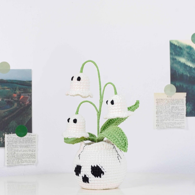 May include: A crocheted white skull-shaped vase holds a plant with three white bell-shaped flowers with black eyes. The plant has green stems and leaves. The vase is on a white surface.