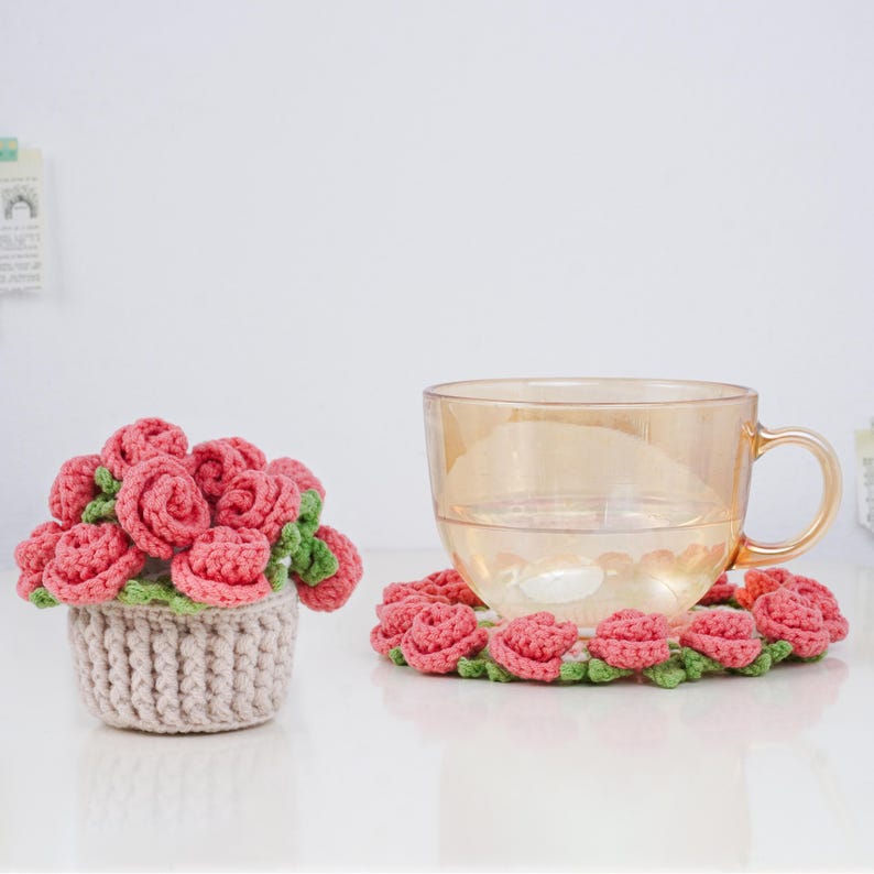 Puede incluir: Un arreglo de rosas de ganchillo en una cesta beige y un posavasos a juego. Las rosas son rosas con hojas verdes. Una taza de cristal se encuentra sobre el posavasos. Art&iacute;culos hechos a mano y decorativos.