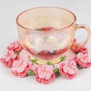Puede incluir: Una taza de t&eacute; de vidrio transparente con asa descansa sobre un posavasos de ganchillo hecho a mano. El posavasos presenta un dise&ntilde;o floral con flores rosas y rosa claro, hojas verdes y un centro amarillo. La taza est&aacute; llena de un l&iacute;quido transparente.