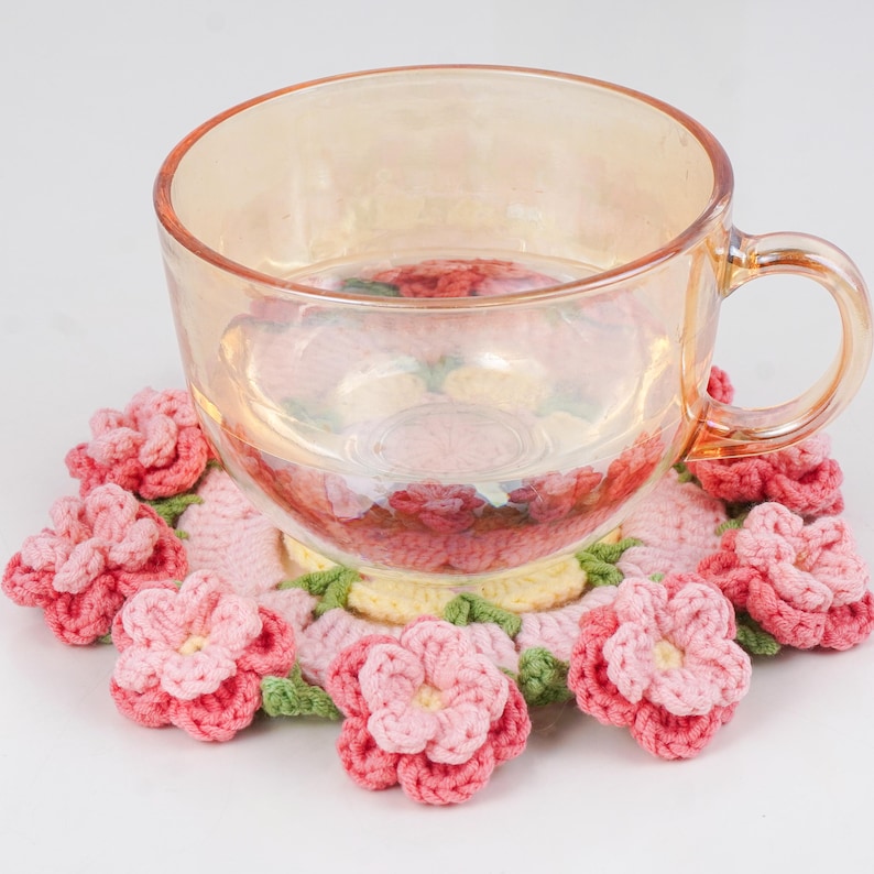Puede incluir: Una taza de t&eacute; de vidrio transparente con asa descansa sobre un posavasos de ganchillo hecho a mano. El posavasos presenta un dise&ntilde;o floral con flores rosas y rosa claro, hojas verdes y un centro amarillo. La taza est&aacute; llena de un l&iacute;quido transparente.