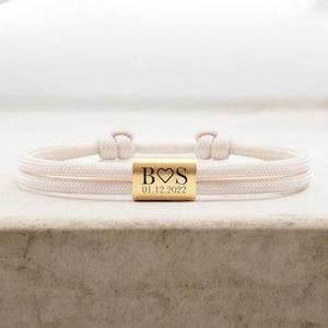 Könnte beinhalten: Ein cremefarbenes Seilarmband mit einem goldfarbenen rechteckigen Perlen. Die Perle ist mit den Buchstaben "B♡S" über dem Datum "01.12.2022" graviert. Das Armband ist an jedem Ende geknotet.