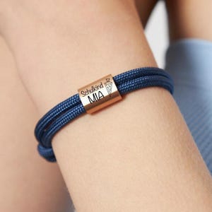 Pode incluir: Uma pulseira azul marinho com uma conta cilíndrica cor de cobre. A conta é gravada com a palavra "Schulkind" acima de "MIA" e um pequeno gráfico. A pulseira é feita de duas mechas de cordão tecido.