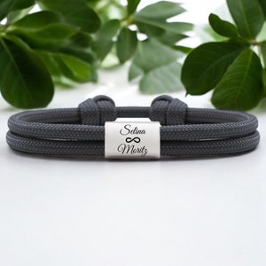 Pode incluir: Um bracelete de paracord cinza de dupla corda com um fecho prateado gravado com os nomes "Selina" e "Moritz" e um símbolo de infinito.