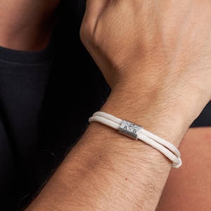 May include: A close-up of a white cord bracelet with a silver cylindrical clasp. The clasp features the letters "A" and "B" with a compass rose design. The bracelet is worn on a person's wrist.