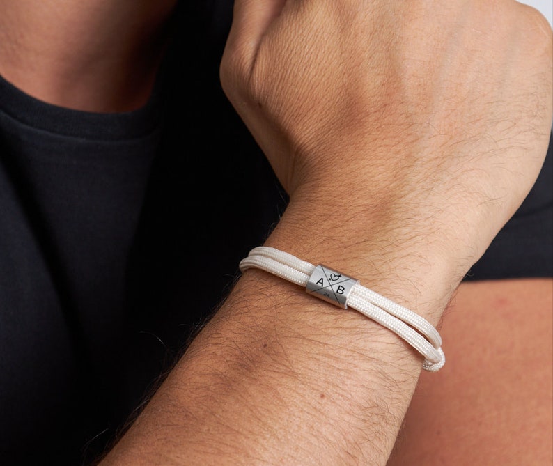 May include: A close-up of a white cord bracelet with a silver cylindrical clasp. The clasp features the letters "A" and "B" with a compass rose design. The bracelet is worn on a person's wrist.
