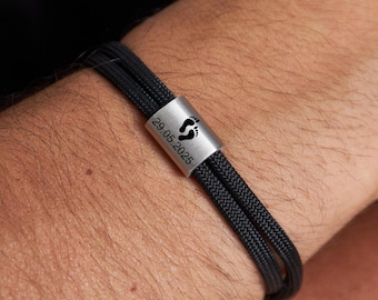 May include: Black braided cord bracelet with a silver-tone metal clasp. The clasp features a footprint symbol and the date "29.05.2025" engraved in black. The bracelet is worn on a wrist.