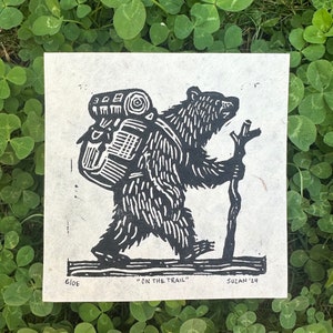 Może przedstawiać: Czarno-biała linorytnica przedstawiająca niedźwiedzia niosącego plecak i idącego z kijem. Druk nosi tytuł "On the Trail" i jest podpisany "Susan '24".