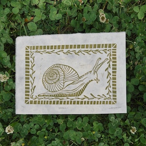 May include: A handmade print on textured paper features a snail illustration in olive green. The snail is centered within a rectangular border with a leafy design. The word "SNAIL" is printed below the image.