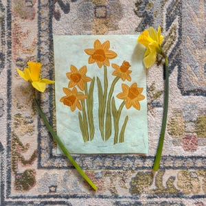 Puede incluir: Naturaleza muerta con una impresión de narcisos naranjas con tallos y hojas verdes sobre un fondo verde claro. Dos narcisos amarillos reales con tallos verdes se colocan a ambos lados de la impresión. La impresión y las flores descansan sobre una alfombra estampada.